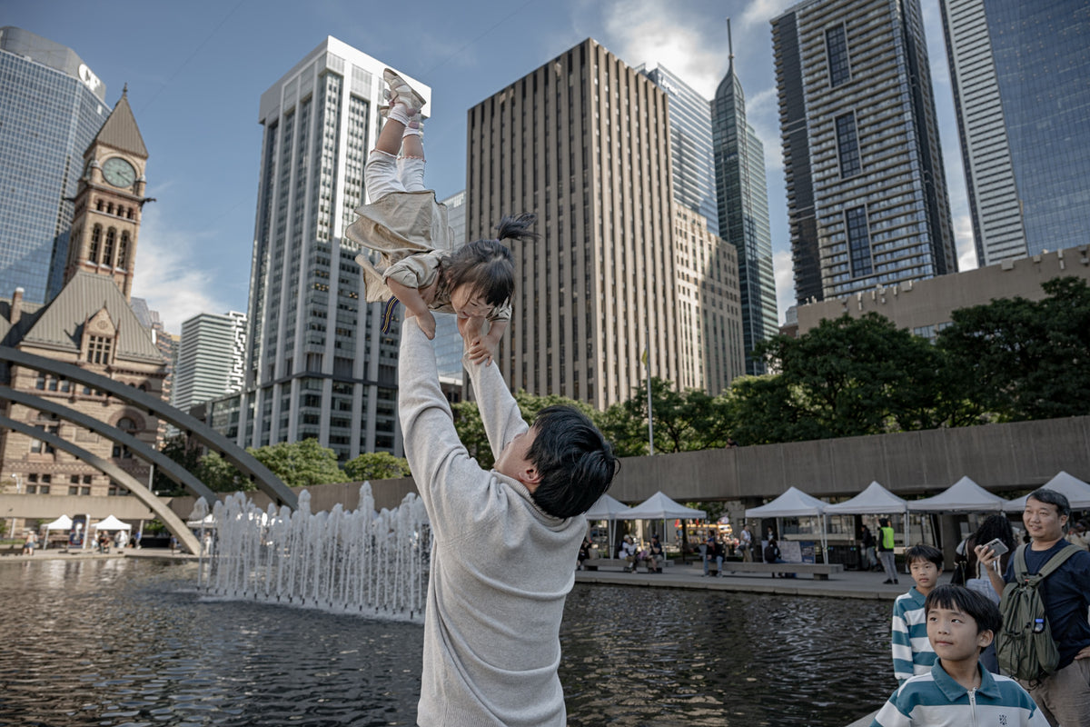 An adult lifts a child beside the fountain and downtown buildings at Nathan Phillips Square, Toronto