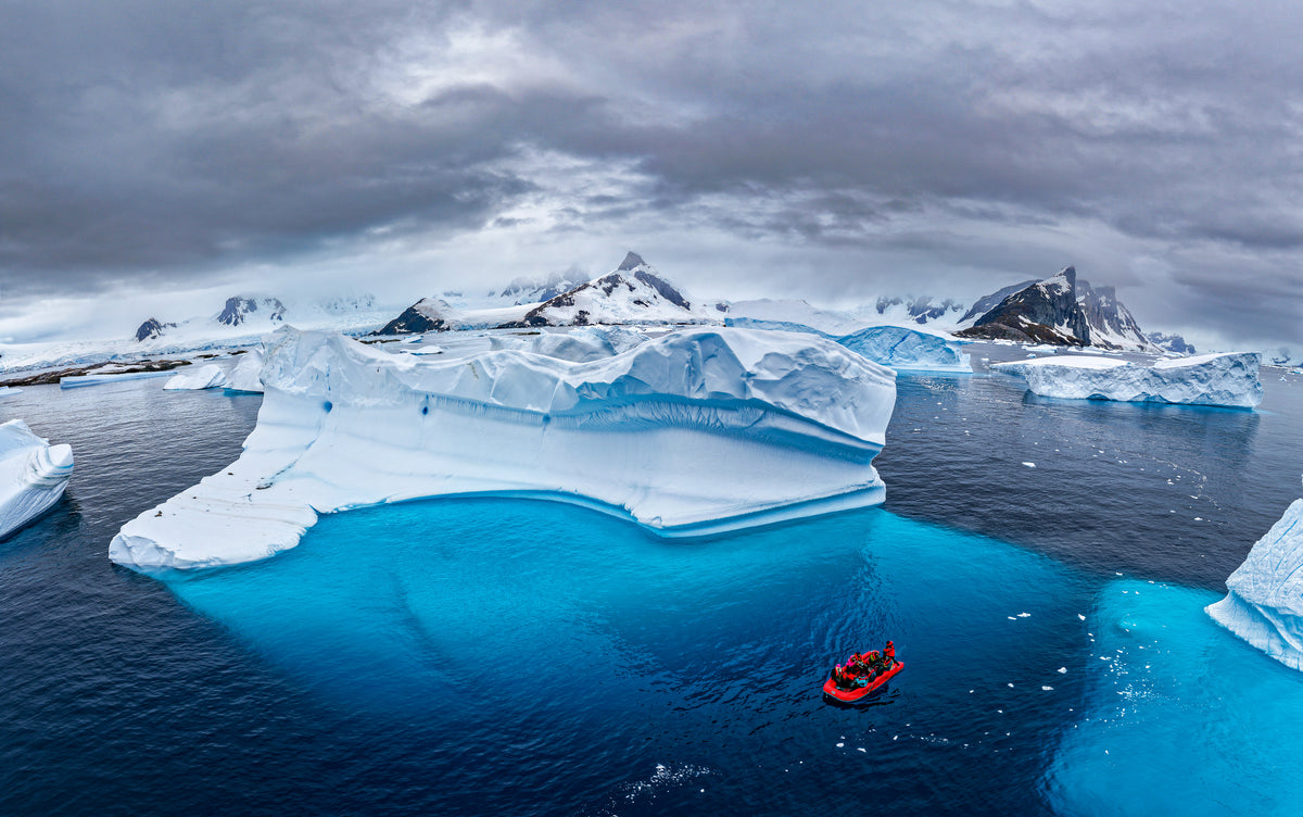 Aerial view of a red Zodiac boat beside blue icebergs and snowy Antarctic mountains
