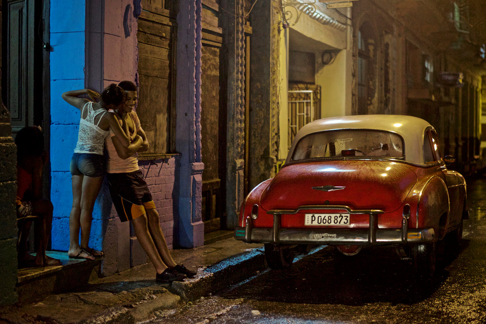 A couple and a red vintage car on a rain-soaked Havana street at night