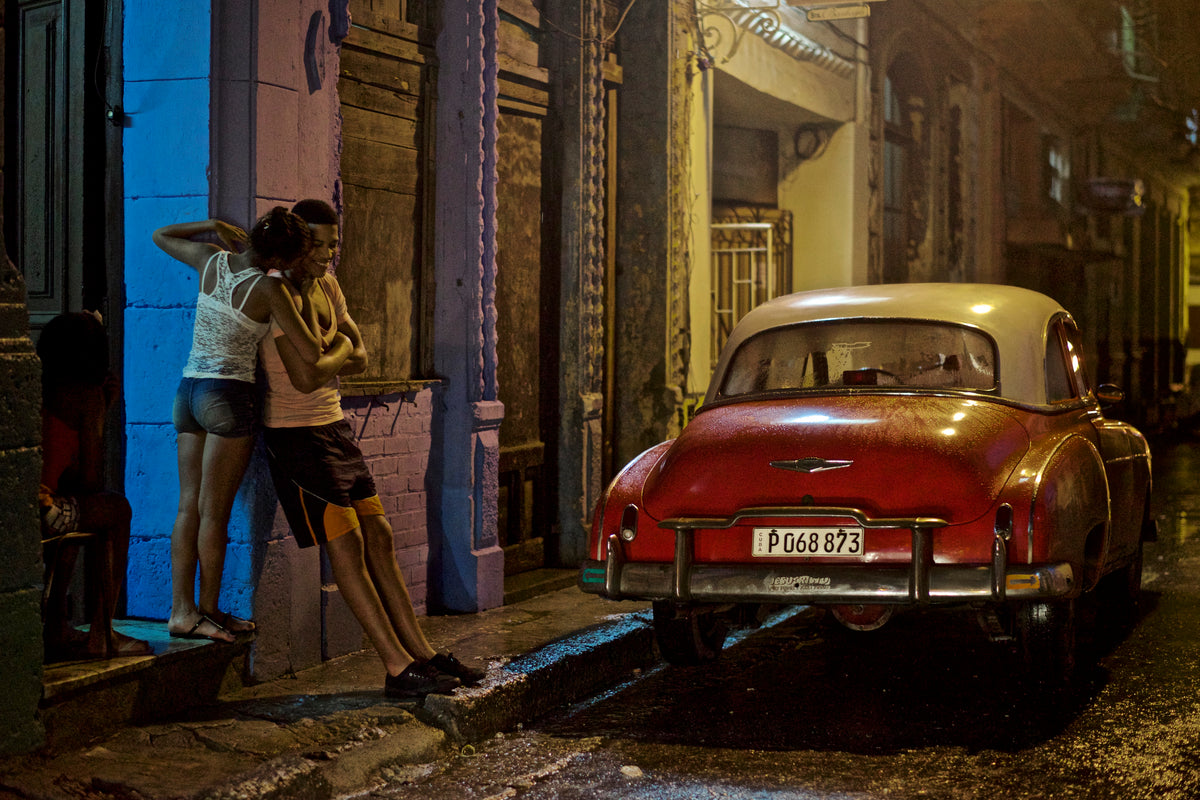 A couple and a red vintage car on a rain-soaked Havana street at night