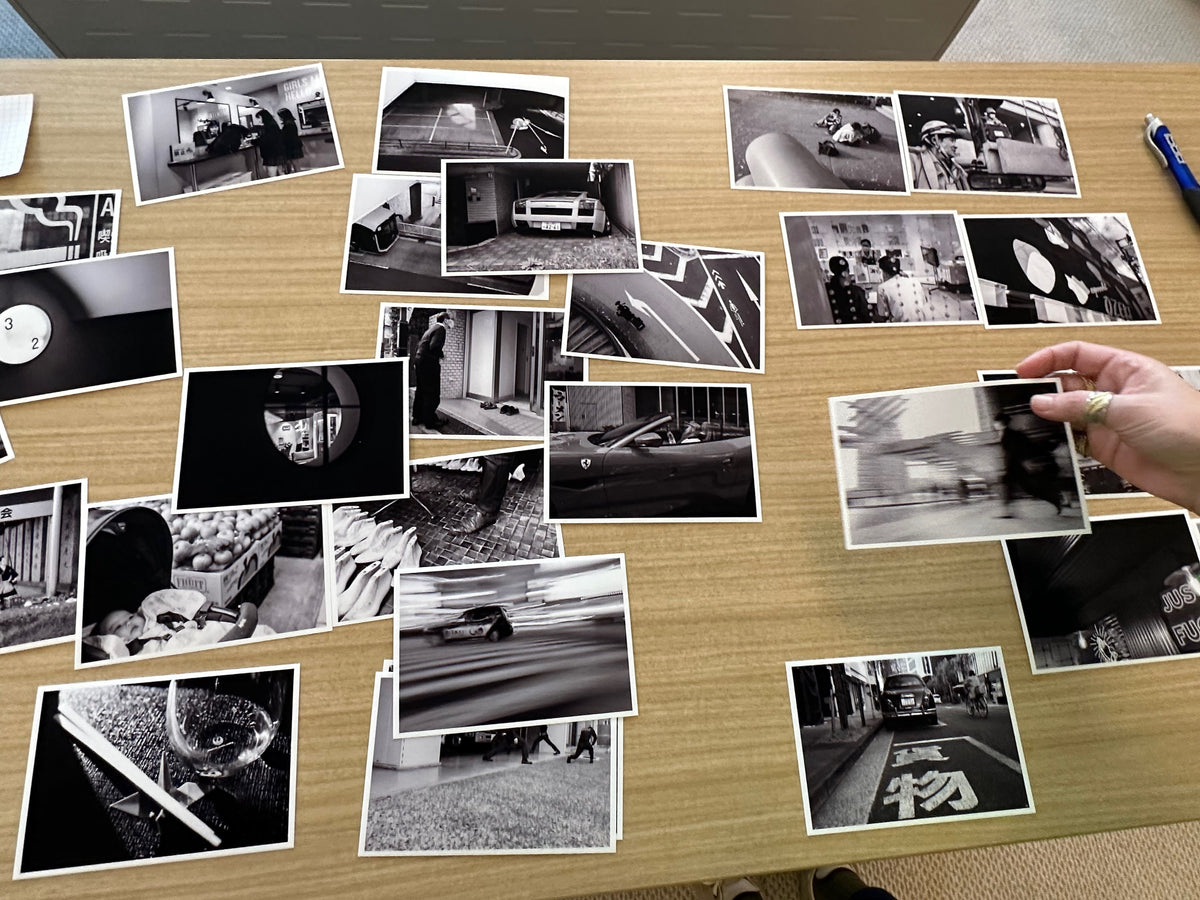 Black-and-white photographic prints spread across a wooden table, with a hand moving one print during an edit.