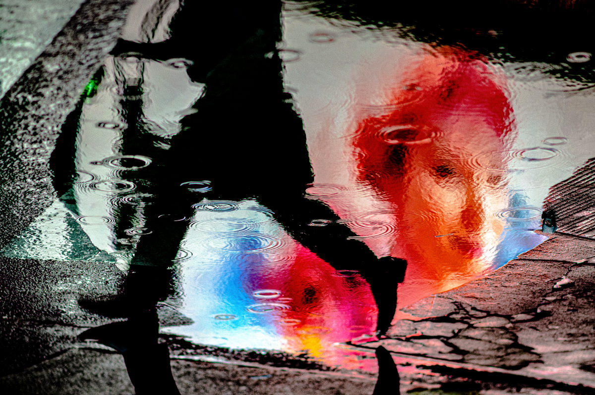 A pedestrian’s silhouette and vivid red and blue reflections in a rain puddle, photographed by Steve Simon